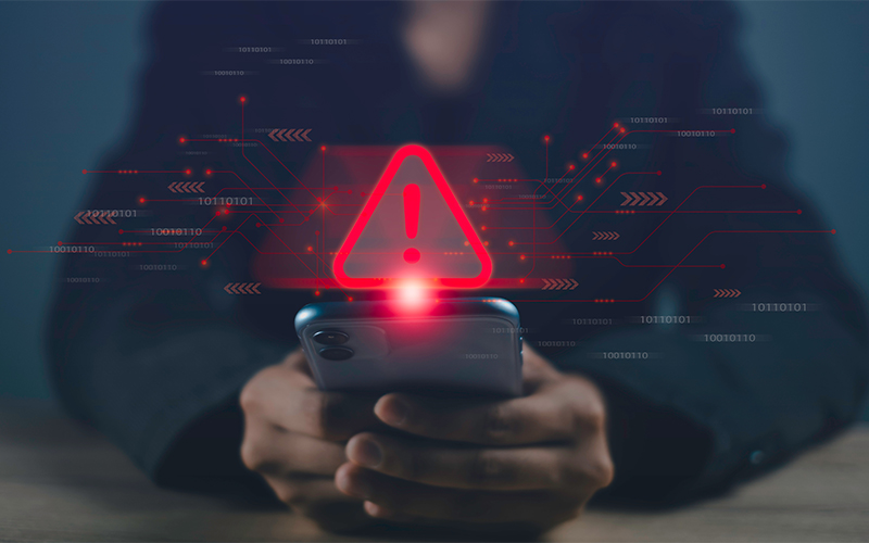 A person holding a smartphone with a red warning triangle and exclamation mark icon above the screen, symbolizing a security alert or error notification.
