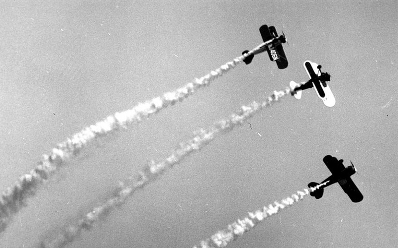 Three biplanes fly in formation through the sky, leaving white smoke trails behind them as they perform an aerial display.