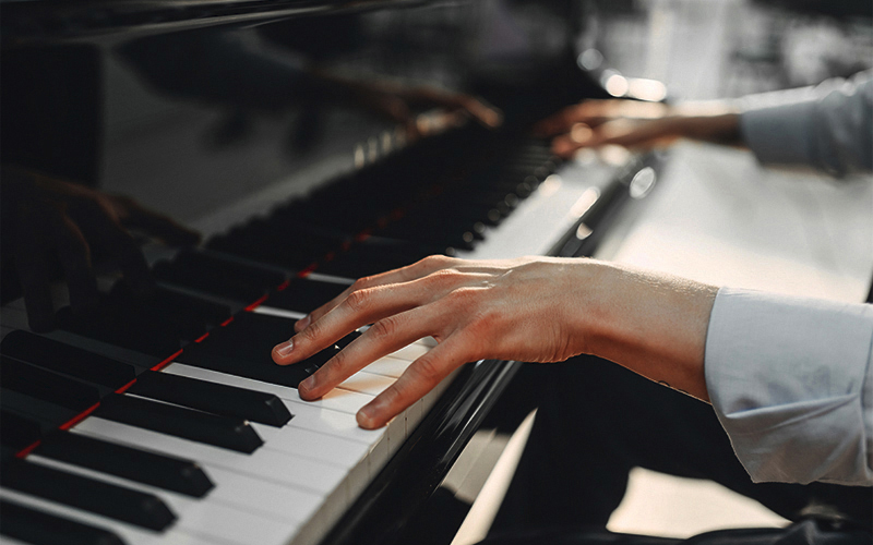 Close-up of a persons hands playing a black grand piano, with sunlight shining on the keys. The person is wearing a light-colored long-sleeve shirt.
