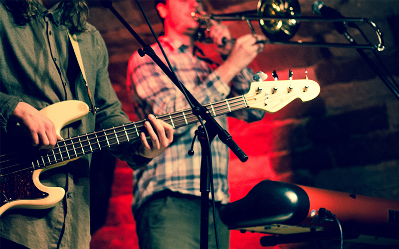 Two musicians performing on stage: one playing an electric bass guitar in the foreground, and another playing a trombone in the background, with warm stage lighting.
