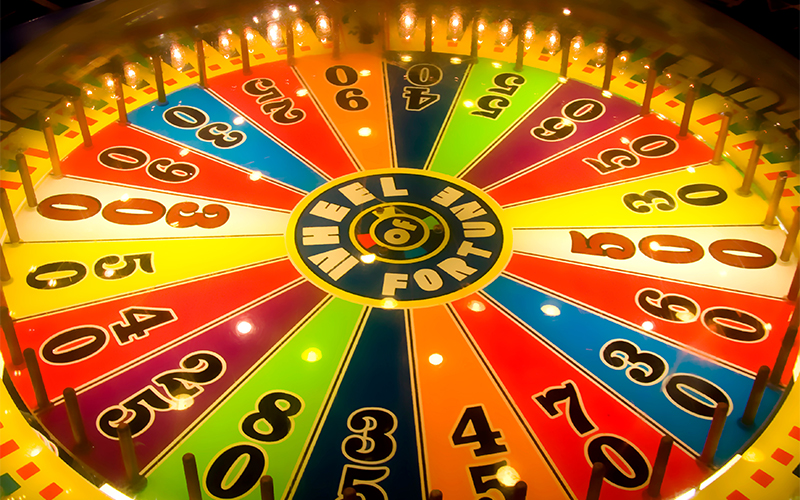A colorful Wheel of Fortune game wheel with numbered sections in various colors, surrounded by lights and pegs, viewed from above.