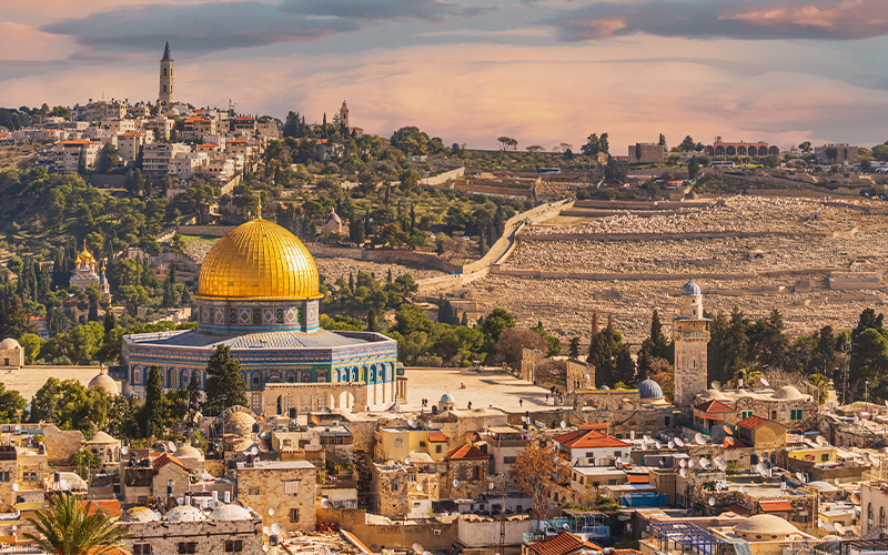 A panoramic view of Jerusalem with the gold Dome of the Rock in the foreground, surrounded by historic stone buildings and hills under a colorful sky.