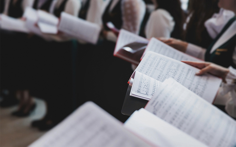 A close-up of people in formal attire holding open sheet music, singing together in a choir. The photo focuses on the music sheets, with blurred singers in the background.