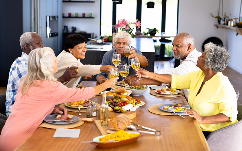 A group of six older adults sits around a dining table, smiling and raising their glasses in a toast. The table is filled with various dishes, salads, and bread in a bright, modern kitchen.