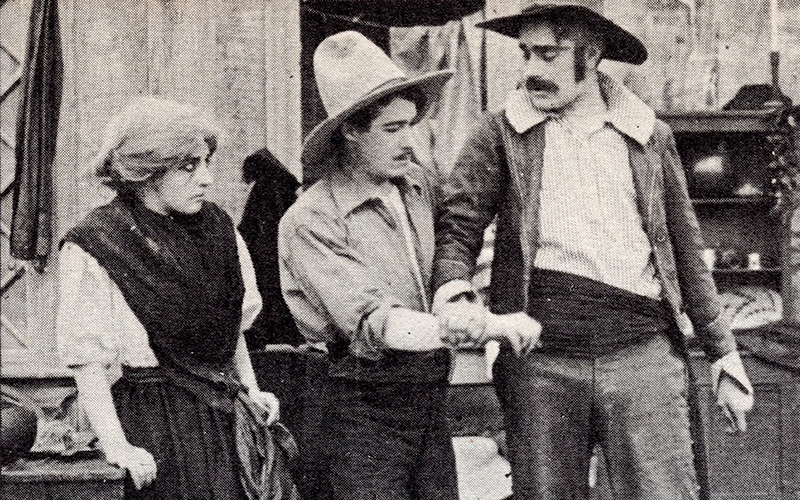 Three people in old-fashioned clothing stand together; a woman looks on as two men, both in hats, hold each others arms in what appears to be a tense or dramatic moment. The scene looks like it is from an old film or play.