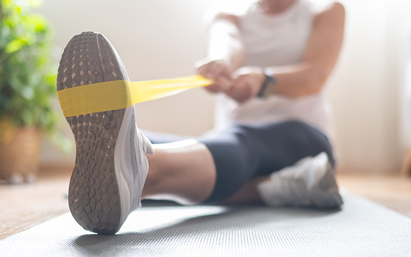 Person seated on a yoga mat stretches their leg using a yellow resistance band, with sunlight coming through a window and a plant visible in the background.