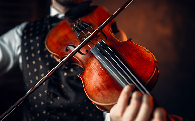 Close-up of a person playing a violin, focusing on the instrument and the musicians hand. The person wears a dark, patterned vest and a bow tie, with a blurred background.