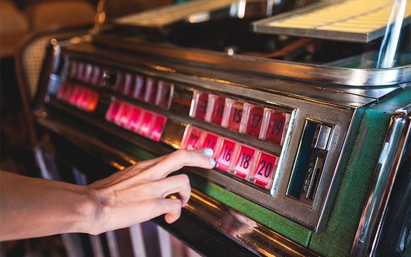 A close-up of a hand selecting a song by pressing a button on a vintage jukebox, with illuminated red numbers visible on the machine.
