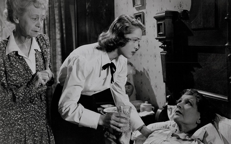 A young woman hands a glass of water to an older woman lying in bed, while another elderly woman stands nearby, watching with concern.