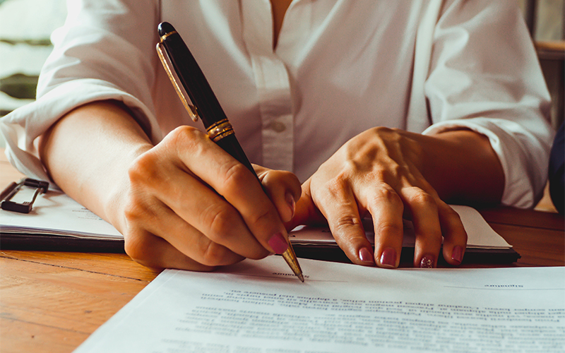 A person in a white shirt is signing a document with a pen at a wooden table. Their other hand holds the paper steady.