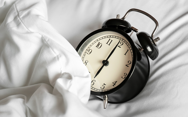 A black retro alarm clock showing 7:00 rests on a white bed with rumpled sheets.