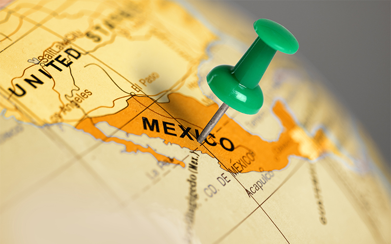 A green pushpin marks the location of Mexico on a yellow and orange globe, with nearby countries and geographic features partially visible.