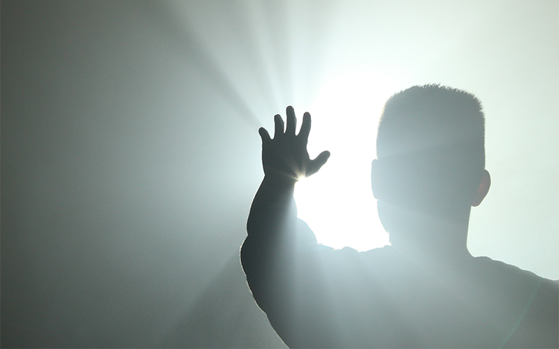 A person stands with their back to the camera, raising a hand toward bright, radiant light shining from in front of them, creating a dramatic silhouette effect.