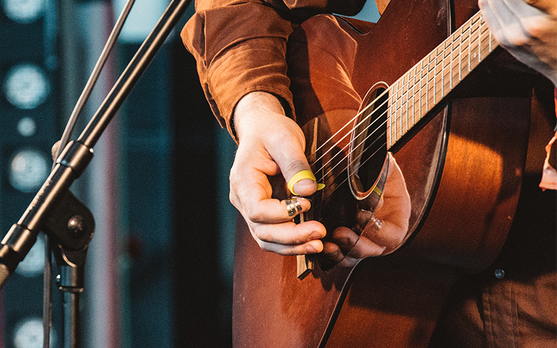 A person wearing a brown shirt plays an acoustic guitar on stage, using a slide on one finger and a pick on their thumb, next to a microphone stand.