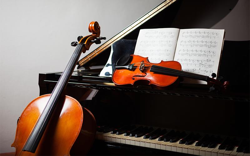 A cello leans against a grand piano, while a violin rests on the piano keys. An open sheet music book sits on the pianos music stand.