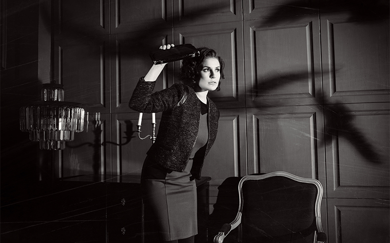 A woman in vintage attire holds a clutch above her head, casting dramatic shadows on a paneled wall, with an ornate chair and chandelier nearby.