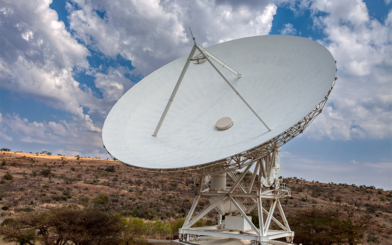 A large white radio telescope dish is positioned outdoors, pointing slightly upward against a partly cloudy sky, with dry grass and scattered bushes in the background.