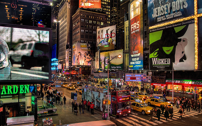 A busy Times Square at night, filled with people, yellow taxis, and bright billboards advertising shows like Wicked and Jersey Boys. Illuminated signs and bustling traffic create a vibrant city atmosphere.
