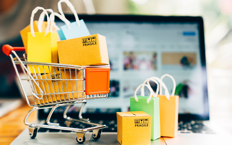 A miniature shopping cart filled with small boxes and colorful shopping bags sits on a laptop keyboard, representing online shopping and e-commerce.