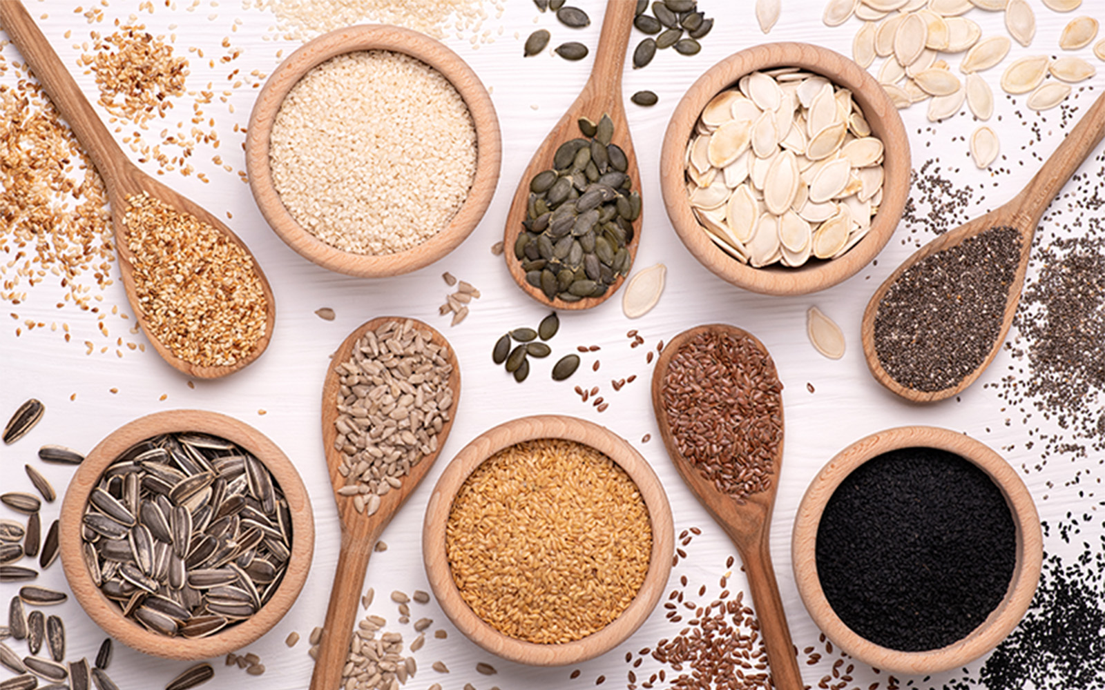 Wooden bowls and spoons filled with various seeds and grains, including sesame, pumpkin, sunflower, flax, and chia, arranged on a white surface.
