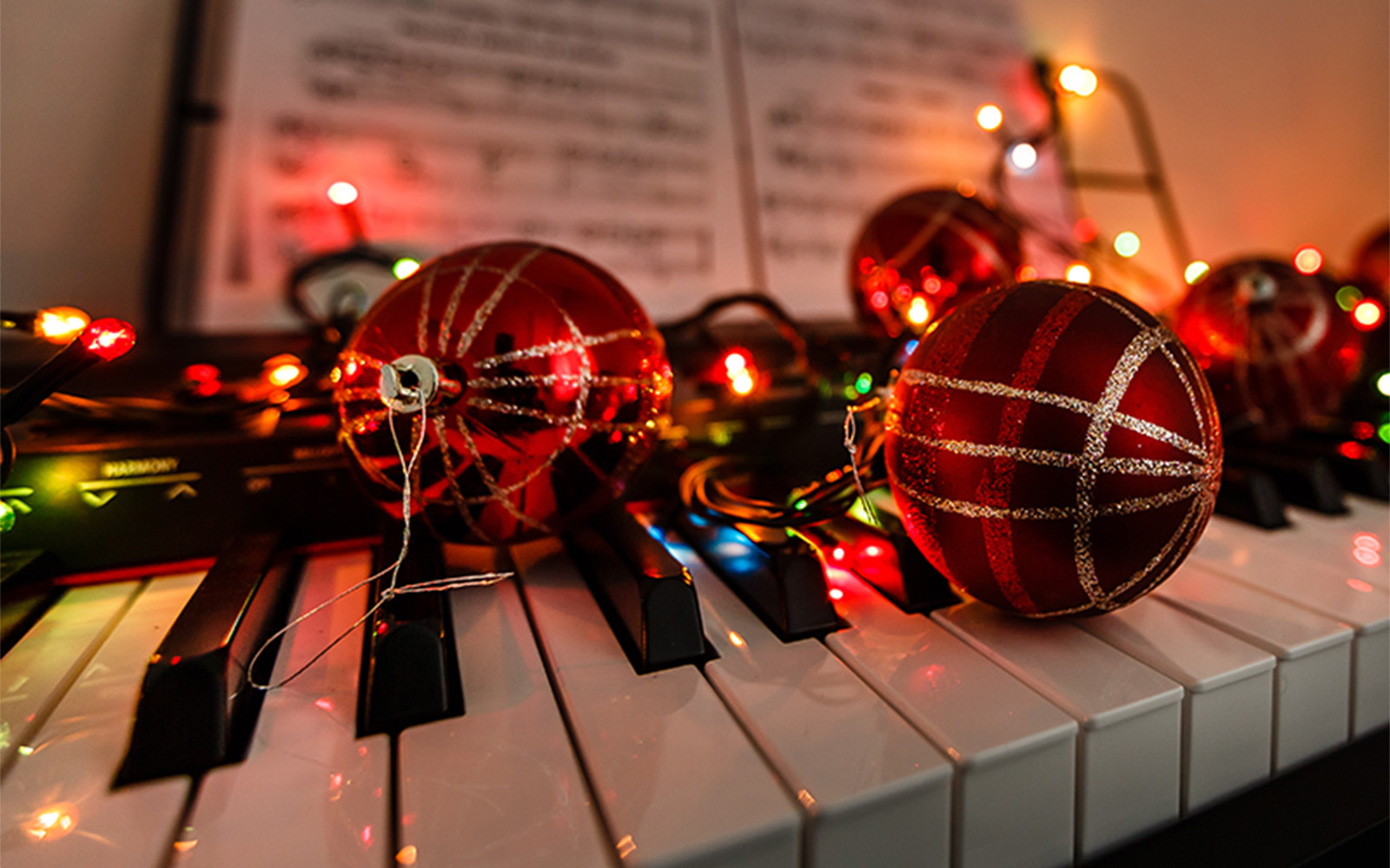 Two red, glitter-patterned Christmas ornaments resting on piano keys, illuminated by colorful fairy lights with sheet music in the background.