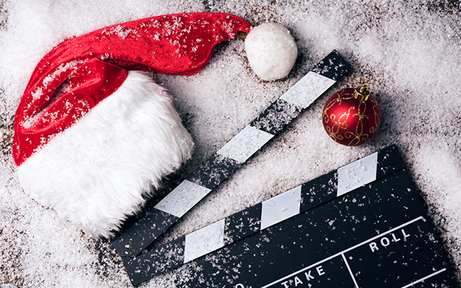 A Santa hat, a red Christmas ornament, and a film clapperboard resting in the snow.